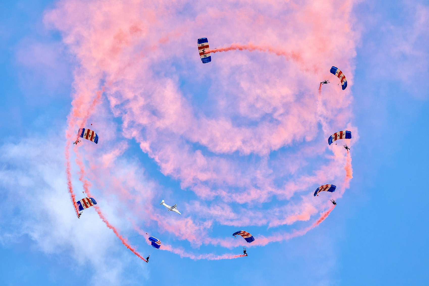 Image shows RAF Falcons Display team parachuting in a spiral, with red smoke trails.
